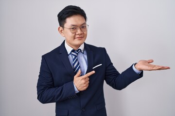 Sticker - Young asian man wearing business suit and tie amazed and smiling to the camera while presenting with hand and pointing with finger.