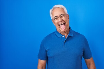 Sticker - Middle age man with grey hair standing over blue background sticking tongue out happy with funny expression. emotion concept.