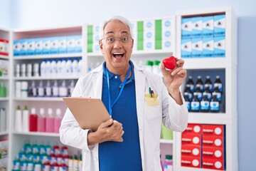 Canvas Print - Middle age man with grey hair working at pharmacy drugstore holding red heart celebrating crazy and amazed for success with open eyes screaming excited.