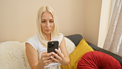 Wall Mural - Relaxed yet serious, a beautiful blonde woman sits at home, engrossed in her online world, texting away on her smartphone in the living room. in her cozy apartment, technology meets solitude.