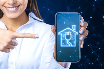 Wall Mural - Happy smiling businesswoman pointing at smartphone with smart home icon on blue polygonal background. Technology and innovation concept.