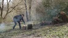 Serious European Man Is Carefully Trying To Chop Small Log With Ax In Awkward Position. Portrait Of Tourist On Vacation Lighting Fire For Grilling. Spring Sunny Forest In Smoke From Cooking.