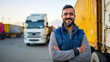 © MP Studio - smiling male truck driver with arms crossed standing in front of a large cargo truck and shipping containers