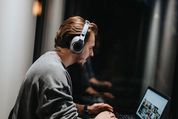 Wall Mural - Concentrated young man with tattoos absorbed in work on his laptop, wearing headphones for focus in a modern workspace setting.