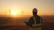 An engineer in a reflective vest and hardhat is inspecting a tablet with wind turbines in the background during sunset.
