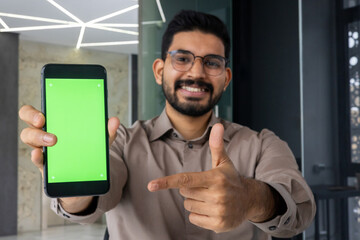 Happy indian male in eyeglasses directing with index finger on mobile phone screen with empty space. Young bearded man recommending new application and approving benefits of installing service.