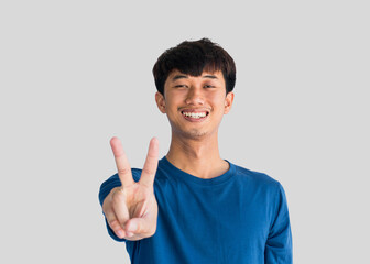 A young Asian man in a blue t-shirt smiling and showing a hand sign second thing isolated on a gray background.