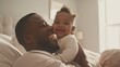 © Wattana - photograph of Happy affectionate young Black dad lifting adorable mixed race baby daughter having fun in bed at home.