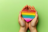 Fototapeta Tęcza - Two hands holding a heart painted like a gay and lesbian flag, isolated on a green background.