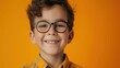 A young boy with curly hair and glasses wearing a yellow shirt smiling against an orange background.