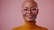 A bald woman with a radiant smile wearing pink glasses and a mustard yellow turtleneck against a soft pink background.
