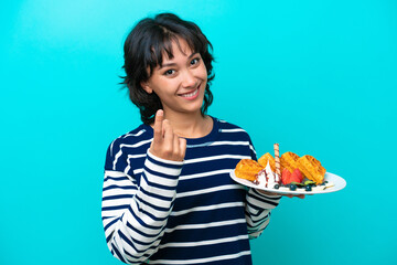 Wall Mural - Young Argentinian woman holding waffles isolated on blue background inviting to come with hand. Happy that you came