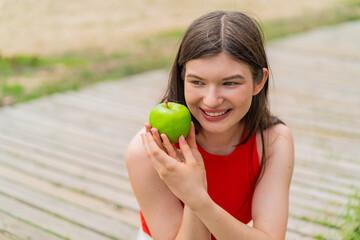 Wall Mural - Young pretty woman at outdoors holding an apple with happy expression