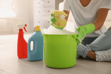 Poster - Woman holding sponge with foam over bucket indoors, closeup. Cleaning supplies