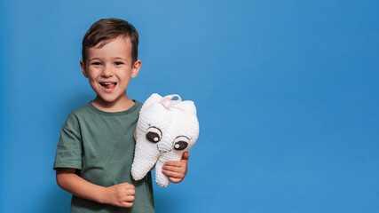 Wall Mural - A smiling boy with healthy teeth holds a toothbrush and a plush tooth on a blue background. Pediatric dentistry. A place for your text. Panoramic photo.