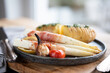 Fried white asparagus wrapped in bacon with herbed potatoes and tomatoes on black ceramic plate and wooden table. Short depth of field.
