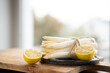 Tied white asparagus with lemon slices on wooden cutting board and black ceramic plate, in front of a kitchen window with shallow depth of field. Kitchen scene for seasonal gastronomy.