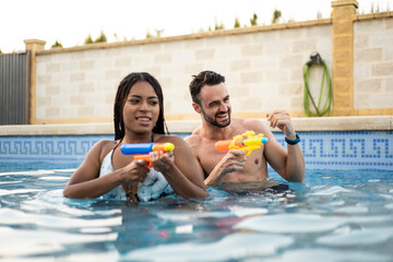 A couple of different ethnicities are playing in the pool with water guns. The young people are shooting with the guns while in the pool. Concept of couples having fun in a pool.