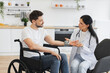 © sofiko14 - Female doctor and patient with disability having talk about diagnosis and medical checkup in modern kitchen. Attentive man with electronic display on hand listening attentively to female physician.