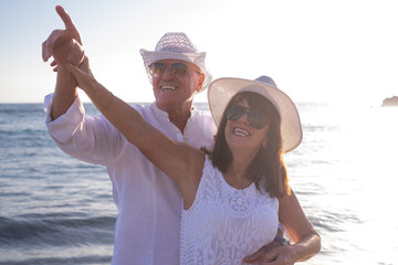 Sticker - Cheerful bonding senior couple hugging on the seashore pointing hand and looking up enjoying vacation and sunset, two smiling people expressing love and tenderness