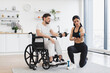 © sofiko14 - Physiotherapist in sportswear showing adult man in wheelchair how do exercises with dumbbells at home. Male patient undergoing rehabilitation after illness or injury in background of modern apartment.