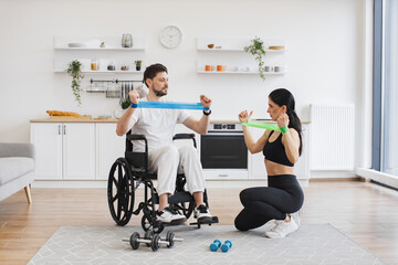 Female physiotherapist or wife showing mature male patient in wheelchair how to exercise with stretching bands at home. Rehab fitness for senior patients concept.