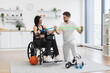 © sofiko14 - Male physiotherapist or husband showing mature female patient in wheelchair how to exercise with stretching bands at home. Rehab fitness for senior patients concept.