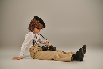 Wall Mural - young photographer in beret and glasses holding camera and sitting on floor, cute boy in suspenders