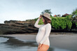 joyful pregnant woman in a snug white bodysuit and a stylish straw hat stands on a sandy beach, holding her hat against the sea breeze, with a soft smile and eyes closed