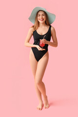 Wall Mural - Pretty young woman in tank swimsuit and hat drinking cocktail on pink background