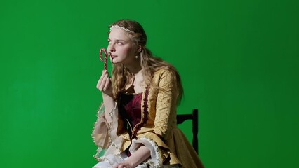 Wall Mural - Woman in ancient outfit on the chroma key green screen background. Female in renaissance style dress sitting holding candy cane eating sweets enjoying.