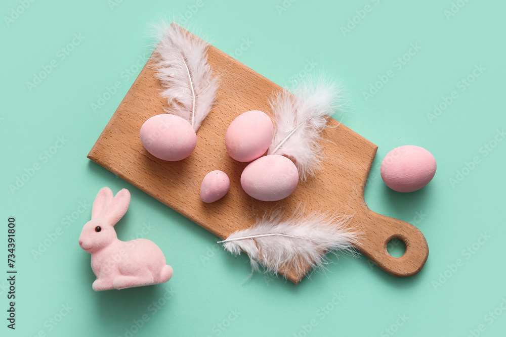 Wooden board with painted Easter eggs, feathers and rabbit on turquoise background