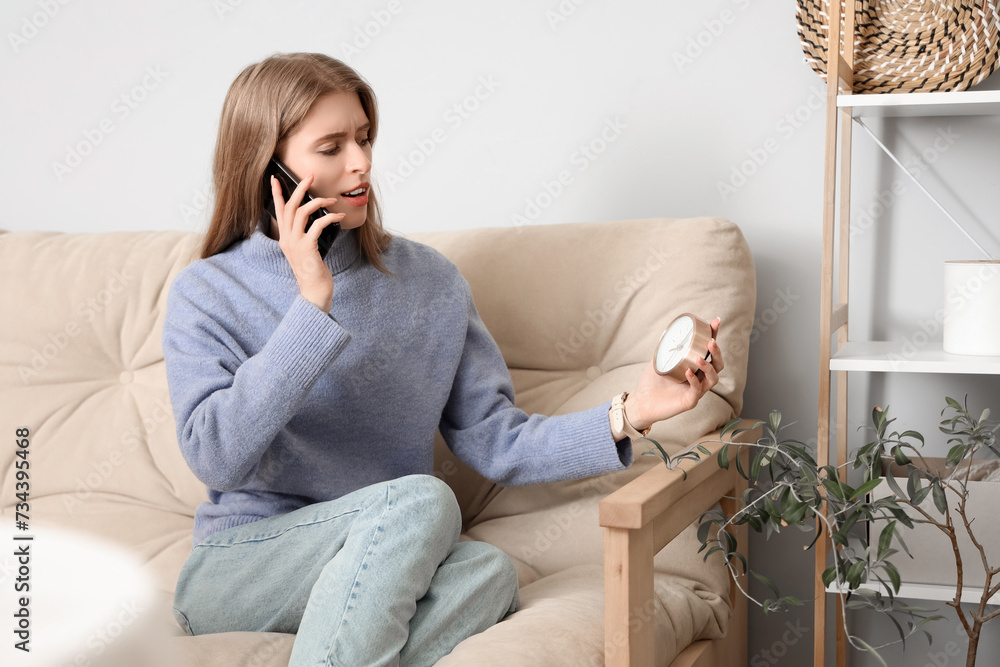 Young woman with alarm clock talking by mobile phone at home