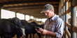 A focused farmer in a white shirt uses a smartphone with a herd of cows in the background inside a barn