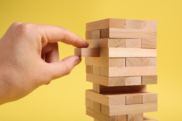 Canvas Print - Playing Jenga. Man removing wooden block from tower on yellow background, closeup