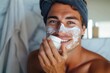 A serene man with a towel wrapped around his head carefully tends to his skin, applying cream in front of a plain wall, his focused expression revealing a sense of self-care and rejuvenation