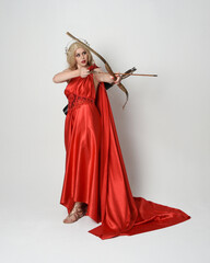 Full length portrait of  blonde model dressed as mythological fantasy goddess in flowing red silk toga gown, crown. Graceful elegant pose  holding  archery weapons, isolated  on studio background