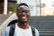 African student wearing glasses and a black backpack stands outdoors near the university appearing happy and successful