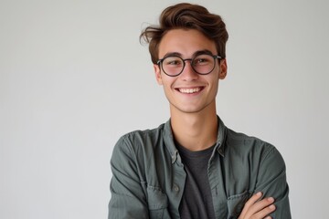Wall Mural - Joyful spectacled young man stands confidently grinning at camera arms folded white backdrop