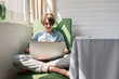 Teenager boy is sitting and holding laptop computer on knees, typing, communicating online, looking at screen, lifestyle home interior, natural bright sunlight from window