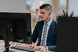 © Maskot - Confident young businessman typing while using computer at desk in office