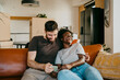 © Maskot - Happy multiracial couple enjoying sharing smart phone while sitting on sofa in living room at home