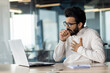 © Tetiana - A young Indian man came down with a virus and a cold. Sitting in the office at the table and coughing, holding his chest from pain and covering his mouth with his hand