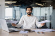 © Tetiana - Financial problems with documents and accounts. Young Indian office worker and businessman sitting at a desk and looking at the camera in frustration, holding papers and spreading his hands