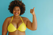 Portrait of happy smiling fat African American woman wearing yellow swimsuit showing thumb up sign isolated on a blue studio background. Travelling, body positive, vacation and summer journey concept.