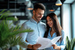 © AI_images - Portrait of two smiling business people man and woman standing in office at their workplace and looking through financial documents. Employees discussing work project or company finances.
