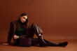 © Victoria Fox - Fashionable confident woman wearing faux leather suit blazer, pants, pointed toe ankle boots, with trendy purple bag, posing on brown background. Full-length studio fashion portrait. Copy, empty space