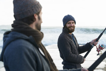 People, fishing and friends at beach for hobby, relaxing and casting a line by ocean with gear. Men, fisherman and together on vacation or holiday, happy and bonding by waves and support on adventure