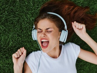 Wall Mural - Beautiful woman in headphones listening to music lying on the grass green lawn in the park in the summer and laughing, happiness in nature, summer lifestyle