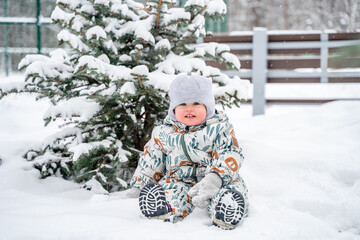 Wall Mural - A child dressed in winter clothes smiles against the background of a snow-covered Christmas tree, it is snowing